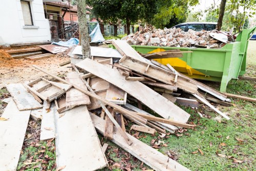 A pile of weathered wooden planks and panels, irregularly stacked on a grassy area in front of a green skip filled with brick rubble and construction debris. The wooden materials vary in length, with some showing signs of age, such as chipped edges, splinters, and faded paint, indicating they are discarded construction or renovation waste. The green skip is situated on a lawn next to a pavement, with trees and residential buildings in the background, suggesting a suburban garden clearance scene. The surrounding environment includes a mix of natural greenery and typical urban outdoor features, emphasizing a typical rubbish removal process in Rotherhithe. The scene appears to be part of a garden or property clearance managed by Garden Clearance Rotherhithe, with natural daylight illuminating the area and highlighting the textures of the waste materials.