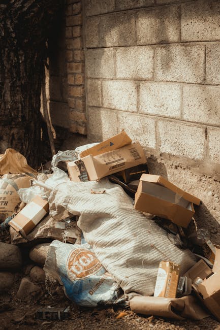 A pile of mixed waste materials and cardboard boxes rests against a weathered concrete block wall outdoors. The debris includes flattened cardboard boxes, some with printed labels and logos, as well a